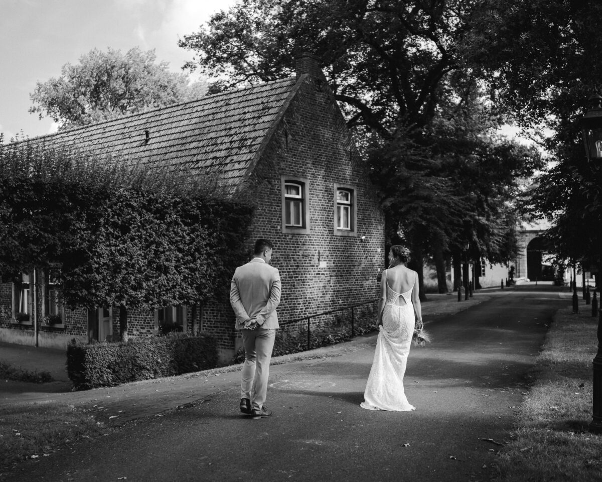 Brautpaar schlendert lässig eine Allee hinauf in Richtung eines großen Tores und lächelt sich an - die Allee ist teilweise von der Sonne durchflutet und beide stehen im Lichtkegel - Paarshooting auf der Hochzeit in Aachen