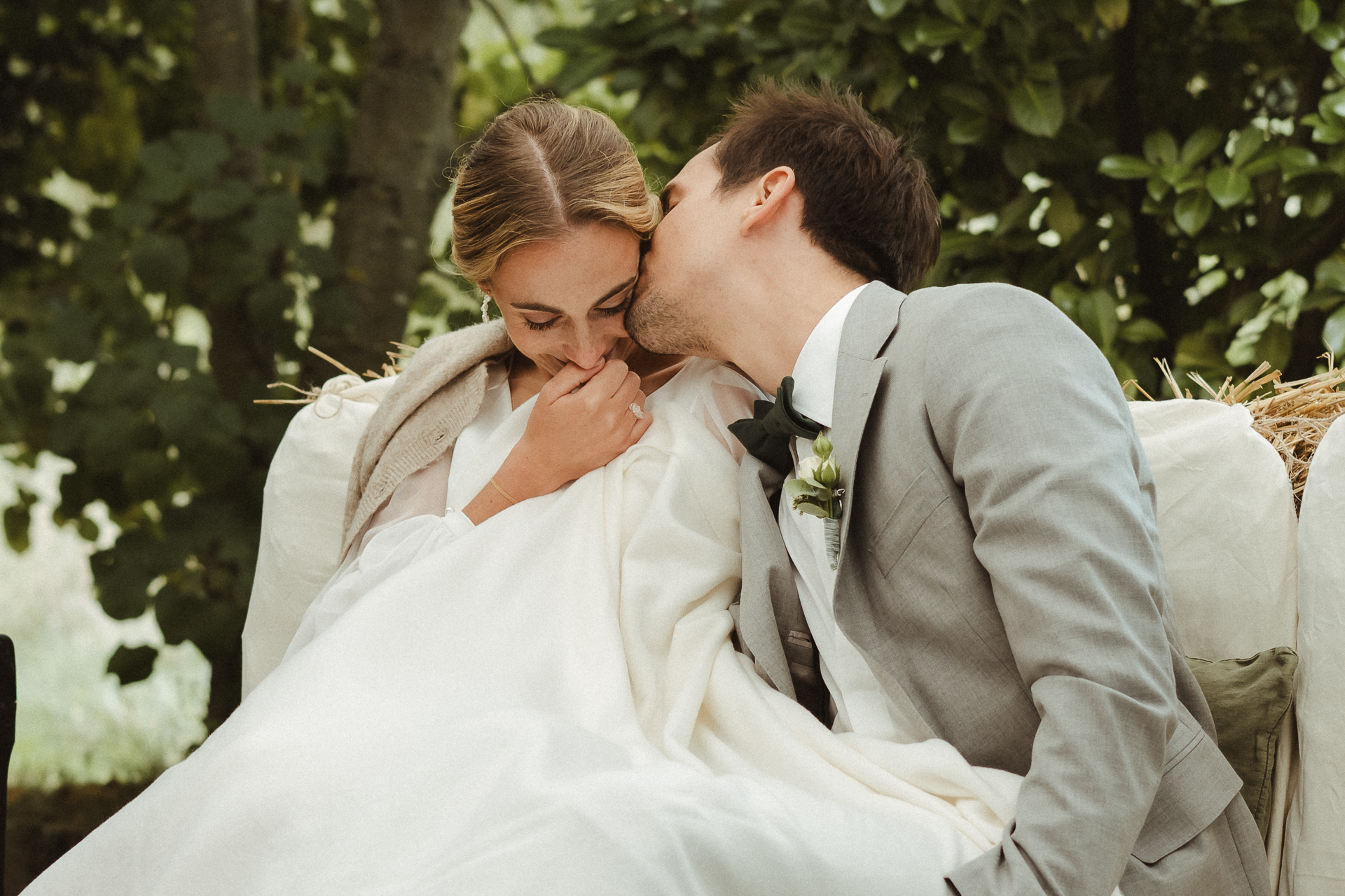 Brautpaar kuschelt unter einer Decke auf einer freien Trauung. Bräutigam küsst seine Braut zärtlich auf die Wange - natürliche Hochzeitsfotos in Düsseldorf
