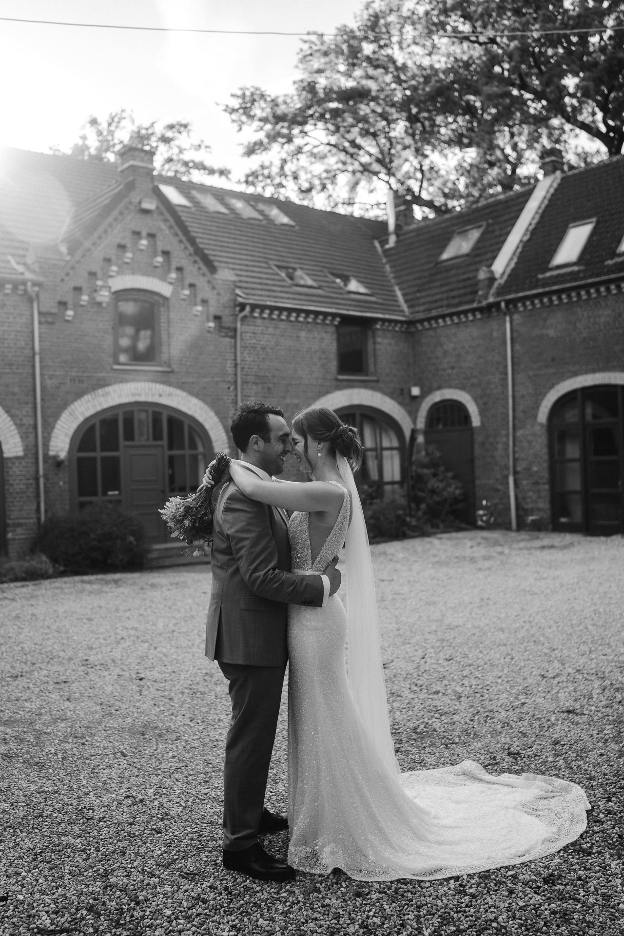 Brautpaar steht im Innenhof ihrer historischen Hochzeitslocation. Das Sonnenlicht wird am Dach des Hauses gebrochen und das Hochzeitspaar steht Stirn an Stirn.