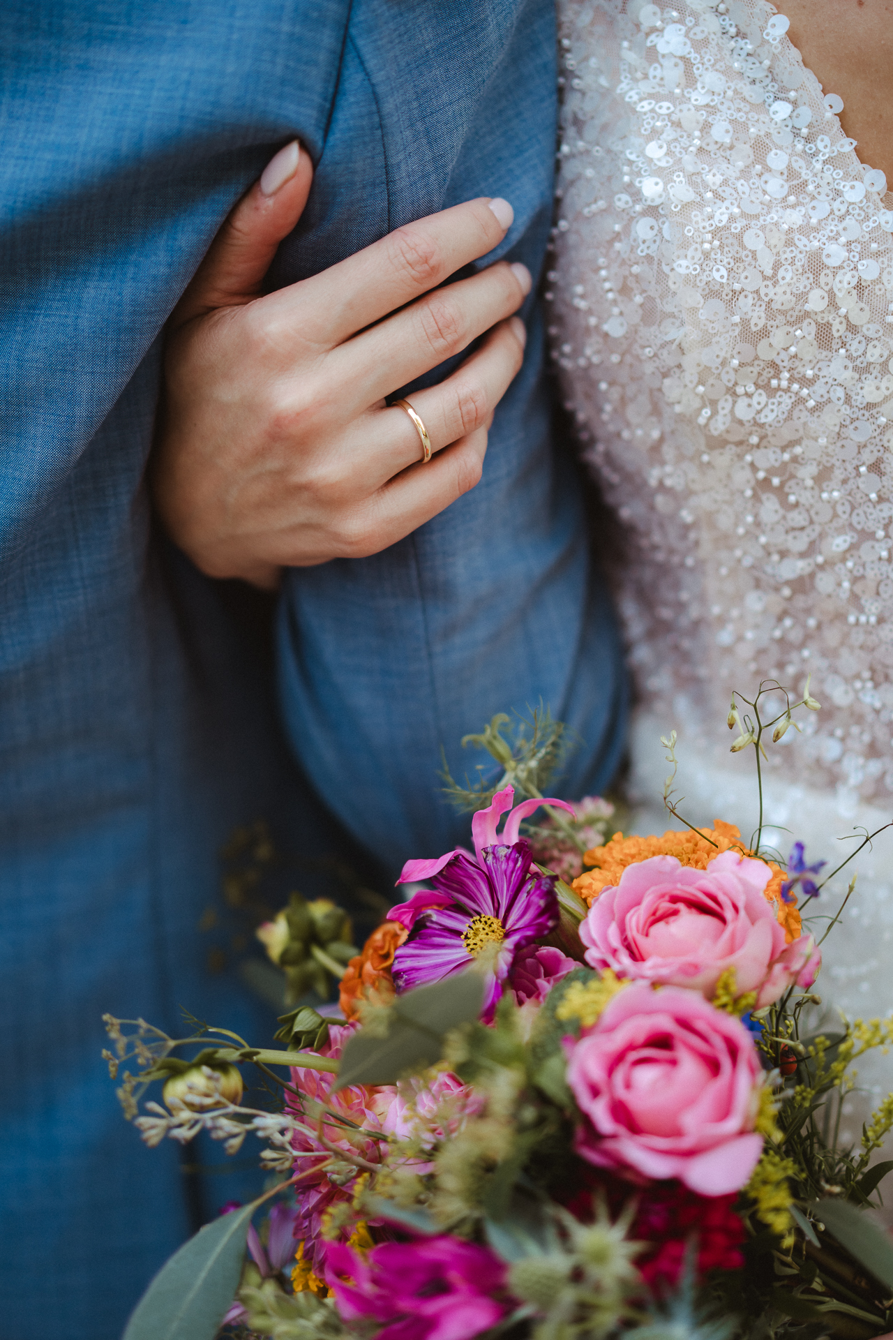 Detailaufnahme von der Hand einer Braut die unter dem Arm ihres Mannes durchgreift. Das Paillettenkleid schimmert und im Vordergrund ist der Brautstrauß zu sehen.