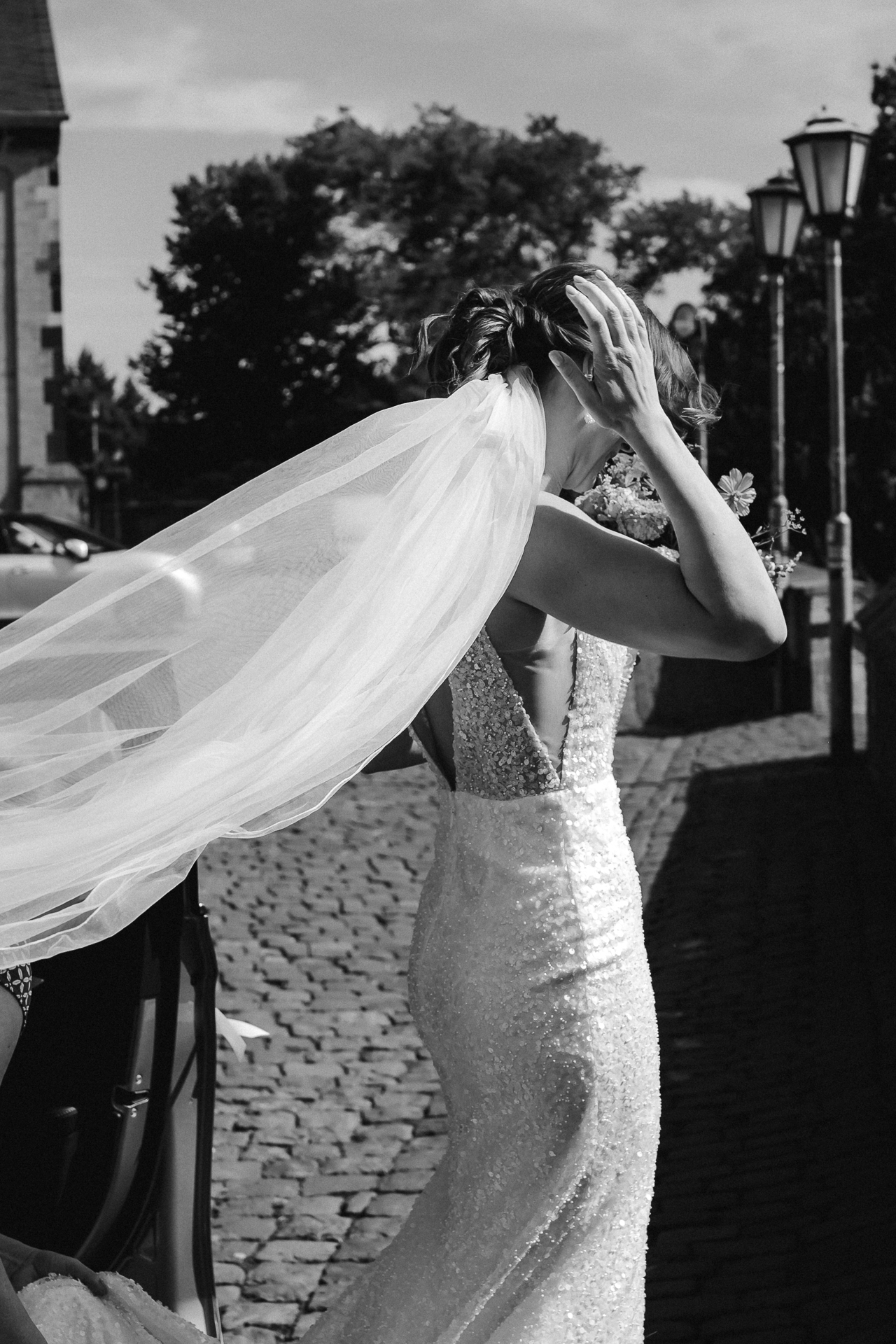 Braut steigt aus dem Brautauto aus. Der Schleier weht im Wind und der Kirchplatz ist im Hintergrund - moderne Hochzeitsfotografie