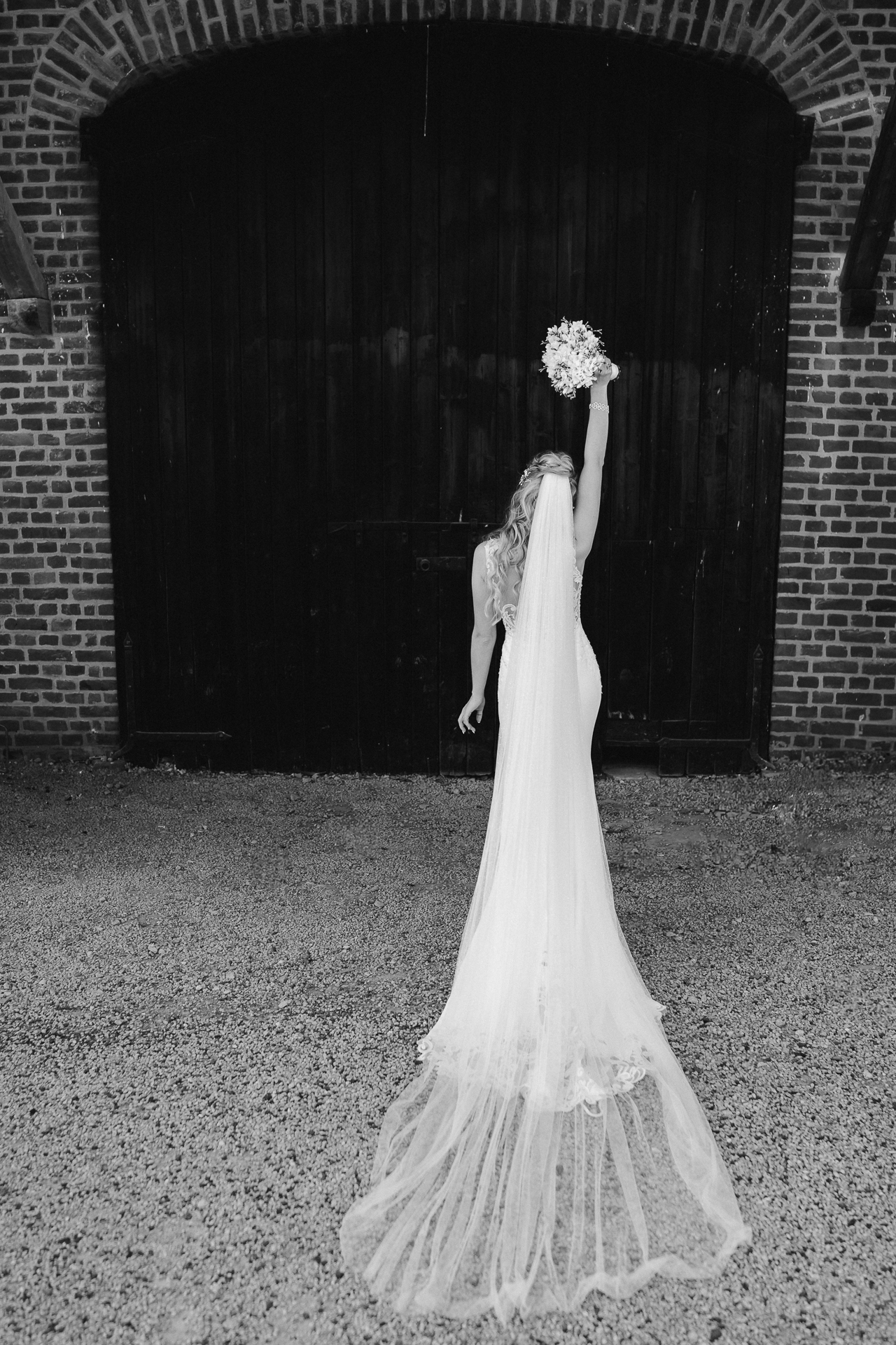Brautfoto streckt ihren Brautstrauß in die Höhe und zieht ihr weißes, traumhaftes Brautkleid mit Schleier hinter sich her - Paarshooting, Hochzeit in Erkelenz