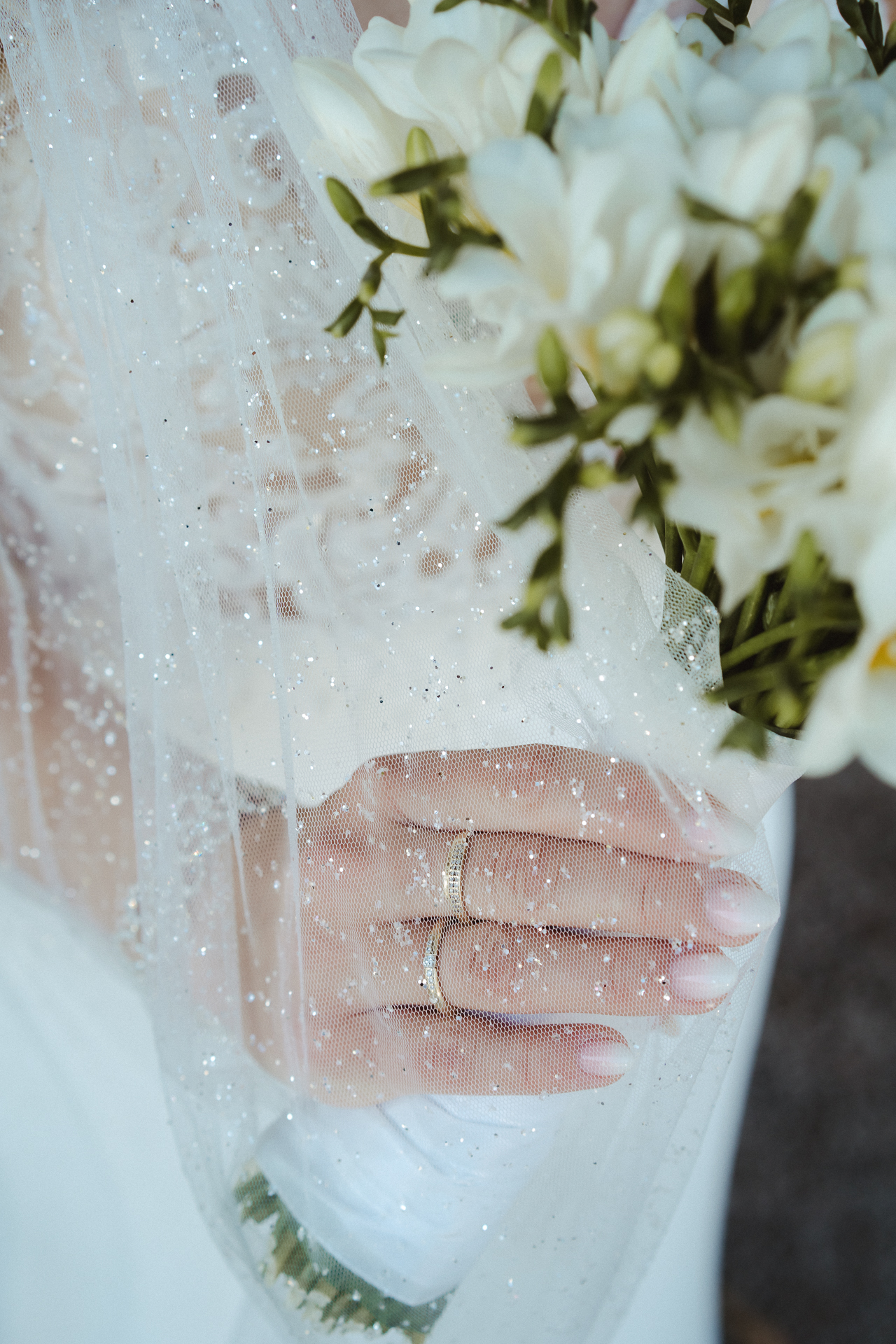 Detailfoto vom glitzernden Schleier der Braut fällt sanft über ihre Hand - an Ihren Fingern trägt Sie Ihren Hochzeitsring und hält ihren Brautstrauß