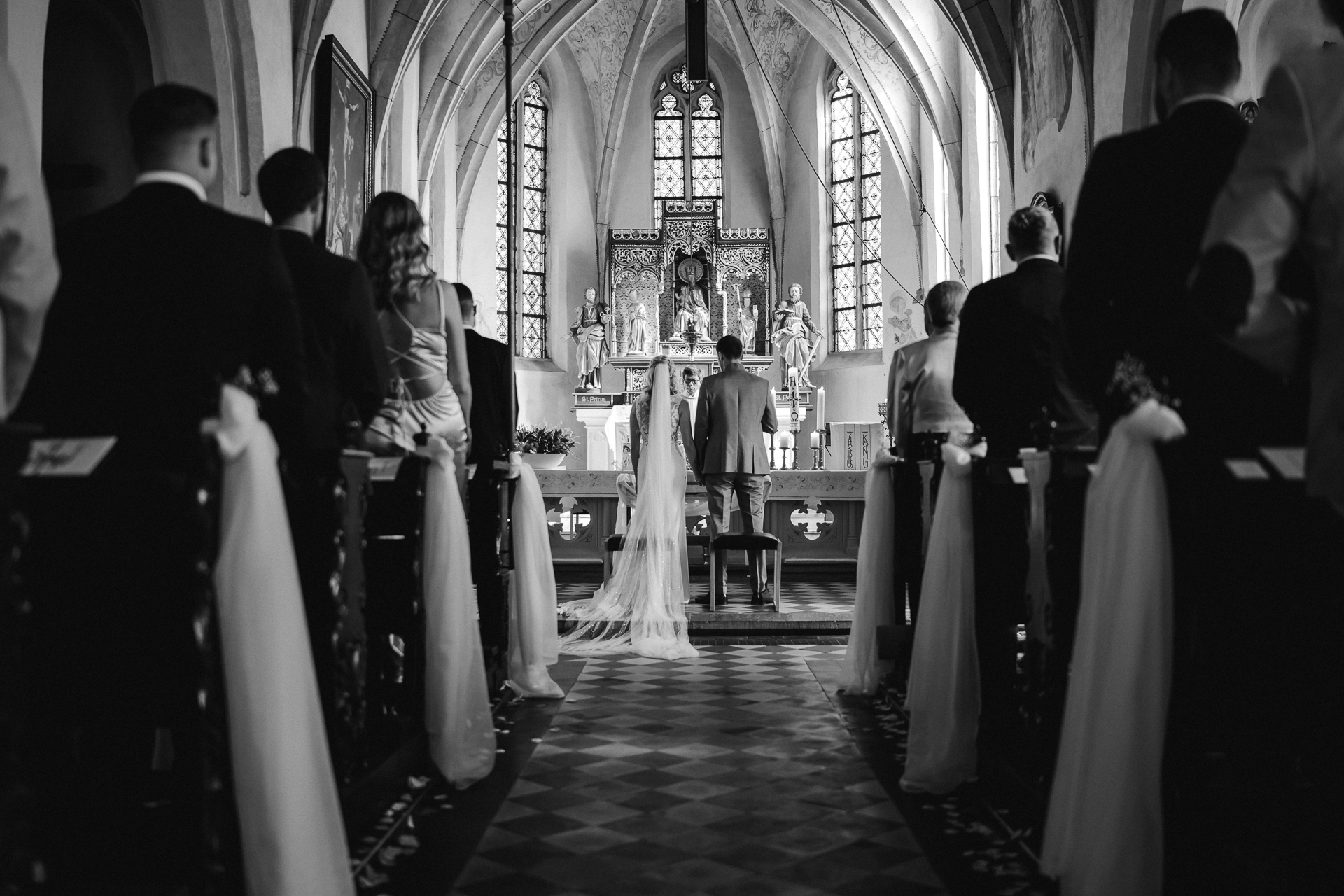 Brautpaar steht Hand in Hand vor dem Hochzeitsaltar. Über ihnen baut sich das Schiff der Kirche auf und links und rechts sind die Sitzreihen mit Gästen zusehen. Das Brautpaar ist im Zentrum des Bildes - kirchliche Trauung in Erkelenz
