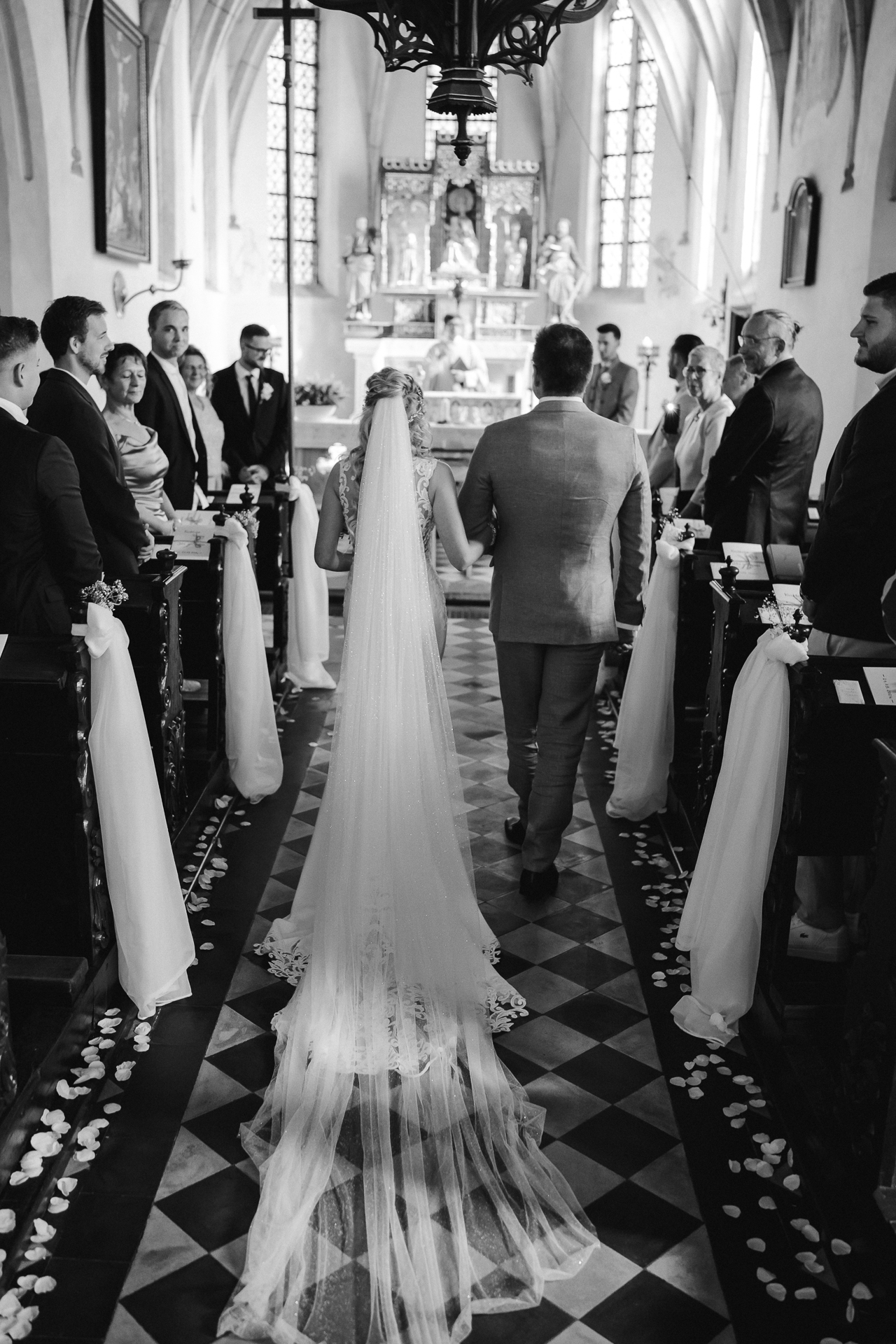 Braut läuft mir Ihrem Brautvater in die Kirche ein. Sie zieht ihren langen Schleier hinter sich her und die Gäste lachen Sie an. Im Hintergrund ist ihr stolzer Ehemann zusenen - Emotionaler Moment in Erkelenz