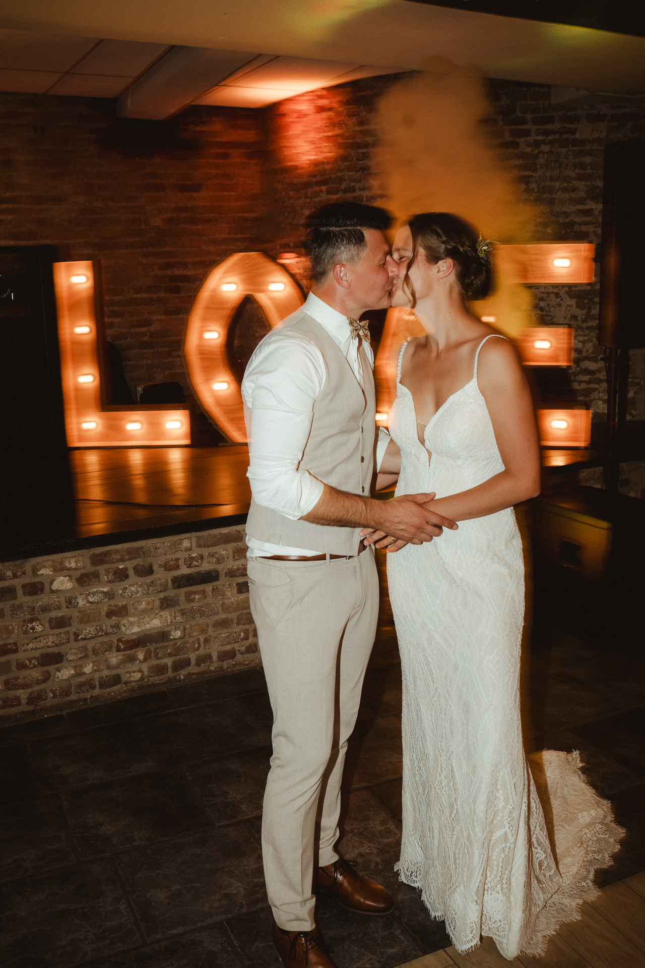 Hochzeitspaar küsst sich vor einem love LED-Schild. Hinter ihnen steigt der Nebel der Party auf - Hochzeitsfotografie